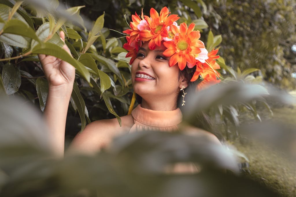 A joyful woman wearing a vibrant flower crown smiles among lush green foliage.
