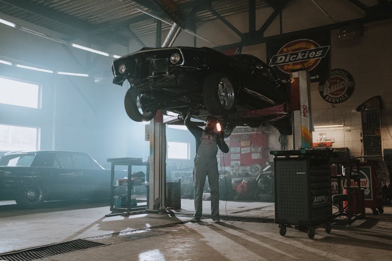 Male Car Mechanic Near Me using a light to inspect a classic car lifted in an automotive garage.allhiphop.co.uk
